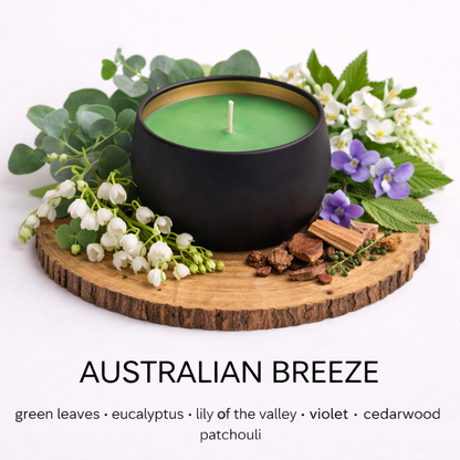 Matte black soy candle with green wax in a gold-lined tin, styled on a wooden base with eucalyptus leaves, lily of the valley, violet flowers, and cedarwood to represent the Australian Breeze scent by Fair Dinkum Scents
