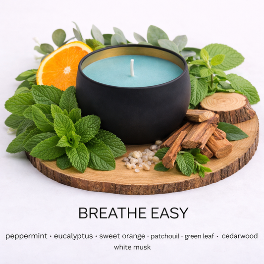Matte black soy candle with blue-green wax in a gold-lined tin, styled on a wooden base with fresh peppermint, eucalyptus leaves, sweet orange, cedarwood, and green foliage to represent the Breathe Easy scent.